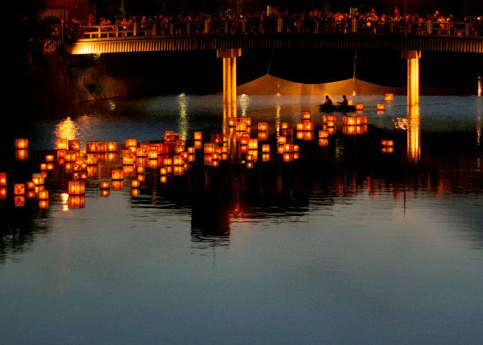 Lanterns floating on a river as onlookers watch from a nearby bridge.